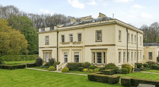 Widworthy Court — a large cream-rendered two-storey house with sash windows, a balustraded balcony and several chimneys, set in manicured gardens