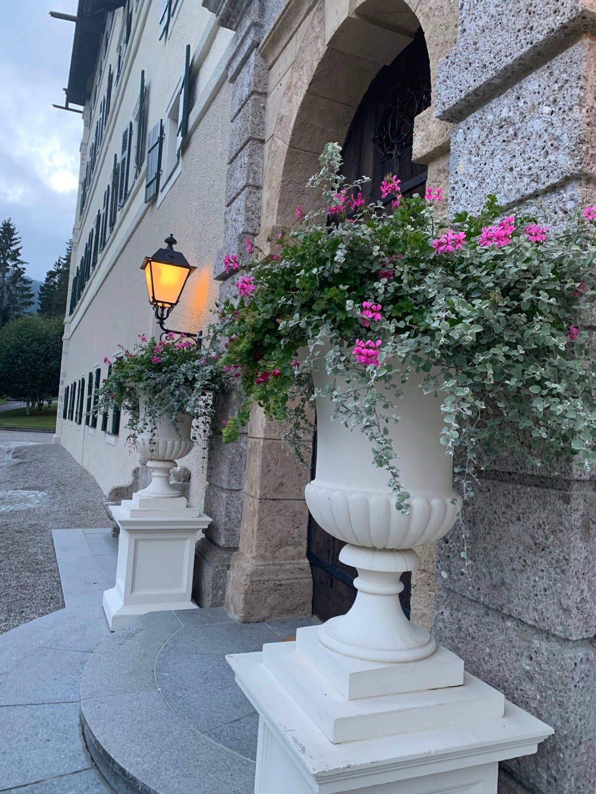 A white stone urn planted with pink and white flowers beside an arched granite doorway and a lantern