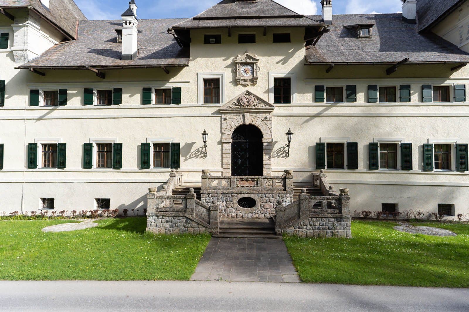 A cream-rendered entrance front with green shutters, a carved stone portal and a double flight of steps