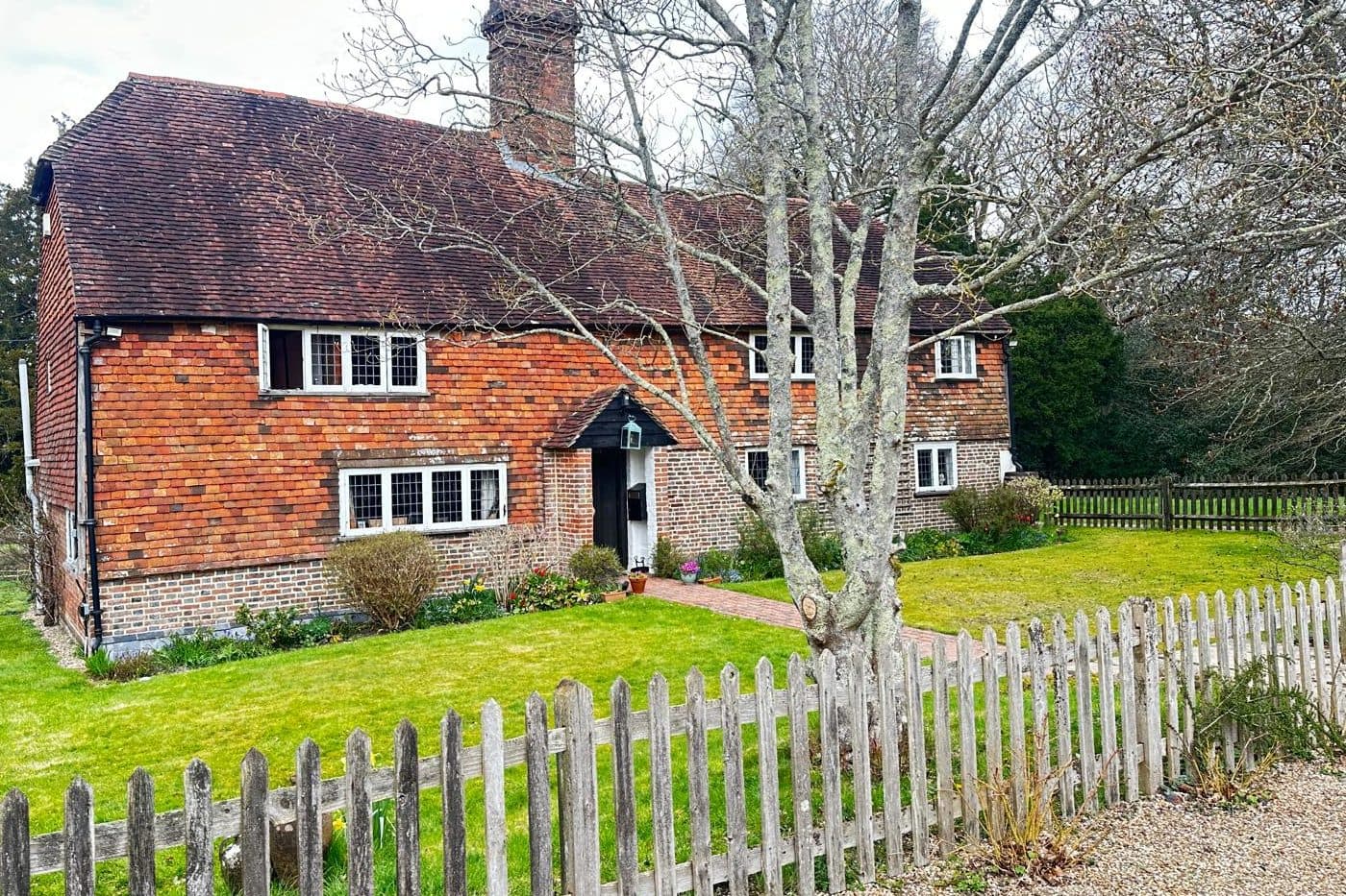 A tile-hung brick farmhouse with a tall chimney behind a timber picket fence and lawn