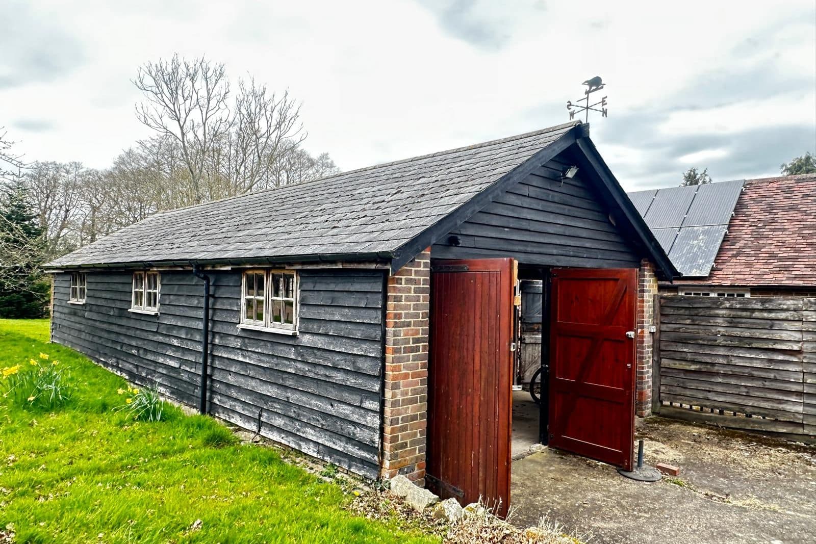 A long, black-stained weatherboarded outbuilding with red-painted doors open, on a lawn