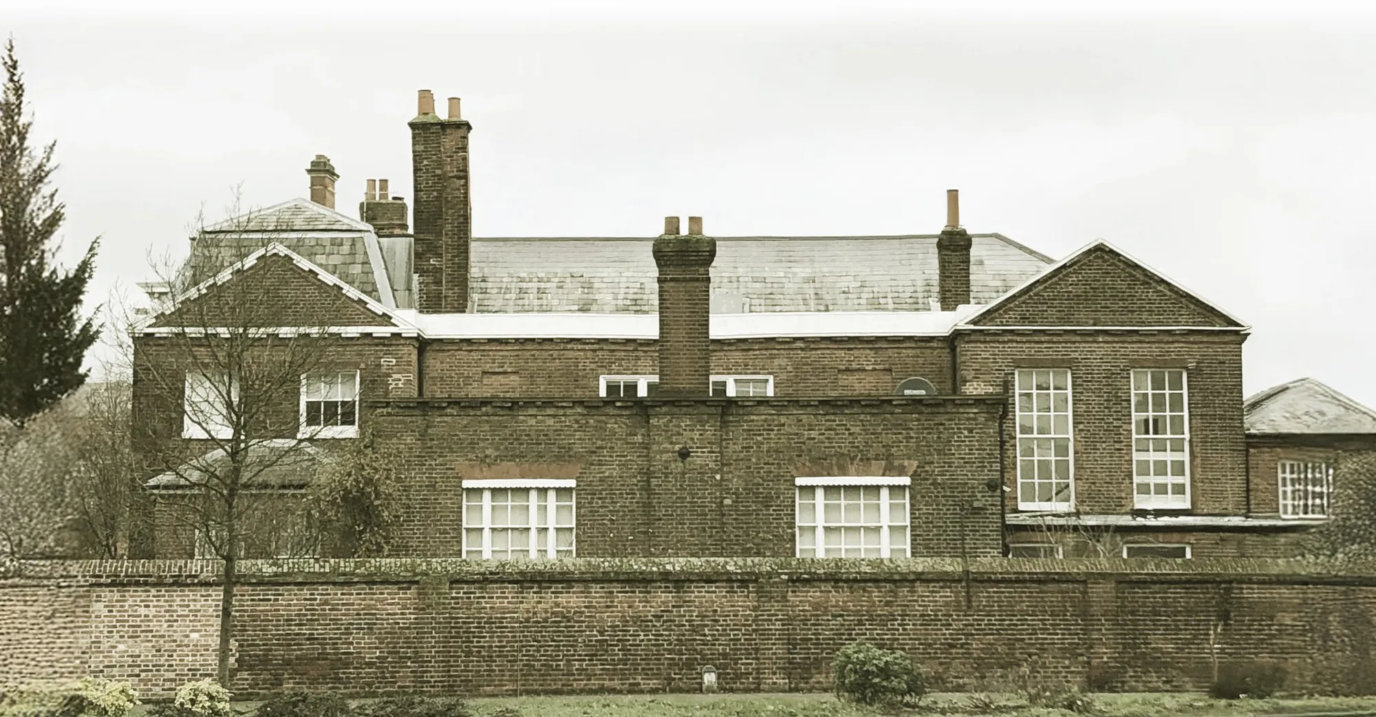 The back of a brick house seen over a garden wall, with slate roofs and several tall chimney stacks above the walls