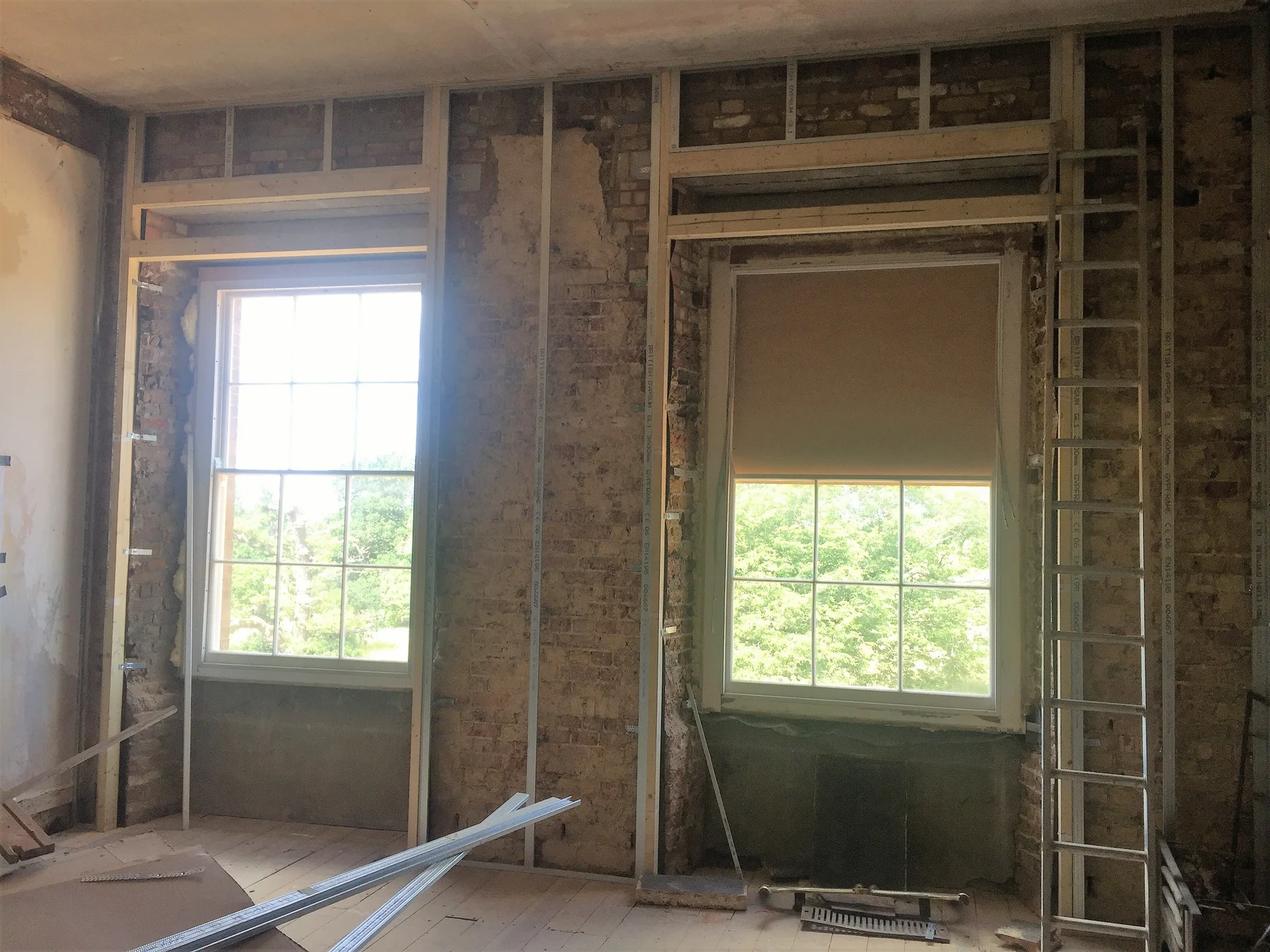 Two tall sash windows in a room with the plaster stripped back to bare brick, timber framing around the openings and a ladder against the wall